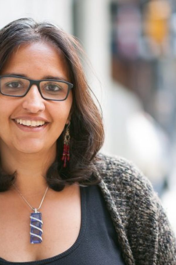 Person wearing glasses and a cardigan smiles at camera against a blurred urban background.