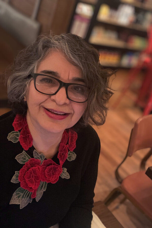 Person wearing glasses and a black top with red floral embroidered neckpiece, smiling at camera in an indoor setting.
