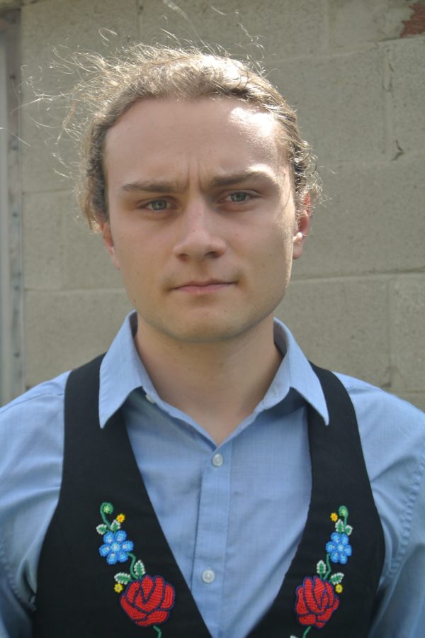 Person wearing light blue shirt and black embroidered vest with floral designs, standing against plain wall background.