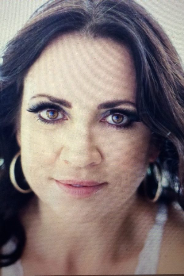 Portrait of a person with shoulder-length dark wavy hair, hoop earrings, and a subtle smile, against a neutral background.