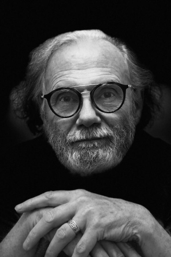 Older person wearing round glasses and a ring, posed against a dark background in black and white portrait photography.