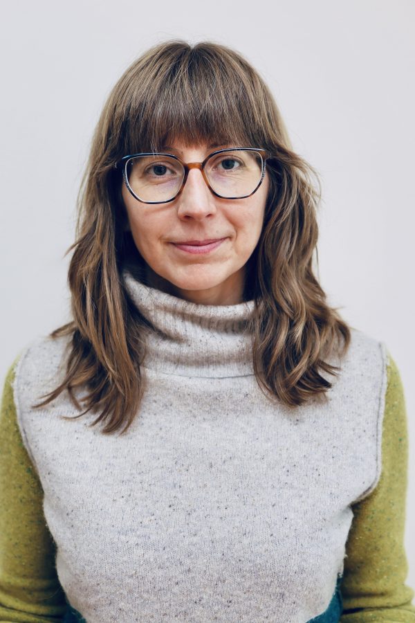 Person wearing glasses and a colorful turtleneck sweater against a white background, smiling at the camera.