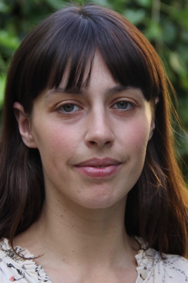 Person with long dark hair and bangs wearing a light patterned shirt, photographed outdoors with green foliage in background.