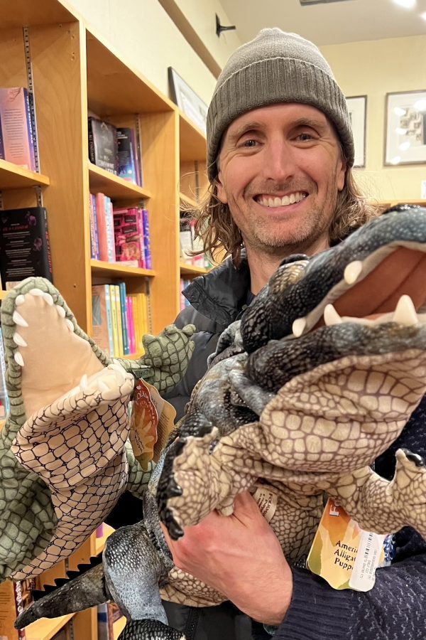 Person holding two reptile figurines in a library with wooden bookshelves in the background.