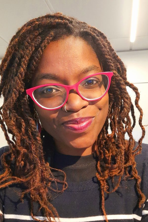 Person with reddish-brown locs wearing bright pink cat-eye glasses and pink lipstick, smiling at camera indoors.