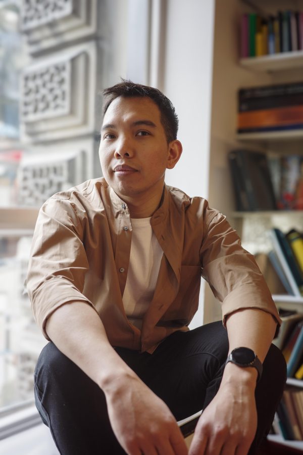 Person in brown shirt sitting by window with bookshelves in background, looking thoughtfully to the side.