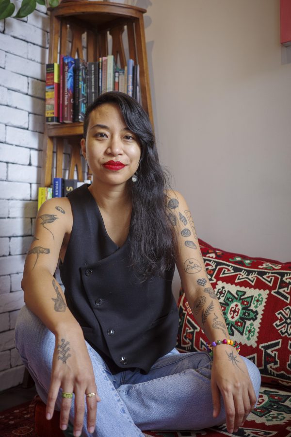 Person with long dark hair and red lipstick wearing a black sleeveless vest, seated indoors with bookshelves and decorative pillows visible behind them.