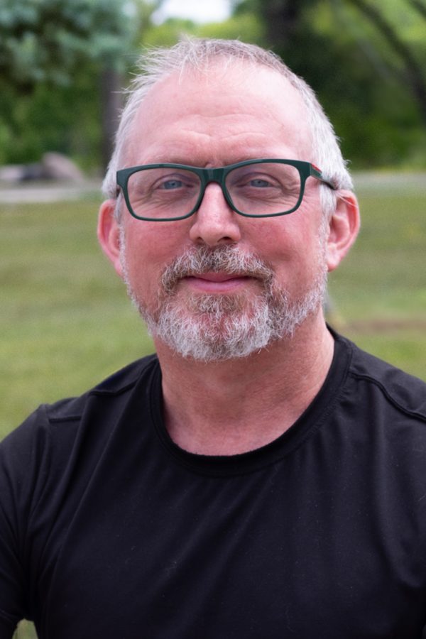 Person wearing glasses and black shirt smiling at camera in a park setting with grass and trees.