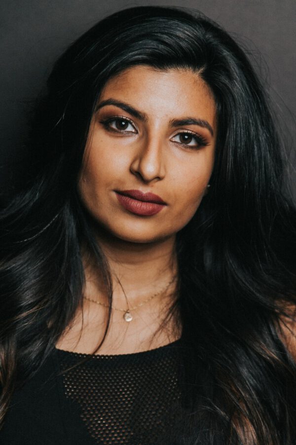 Person with long black hair and warm makeup wearing a black mesh top against a dark gray background, looking toward the camera.