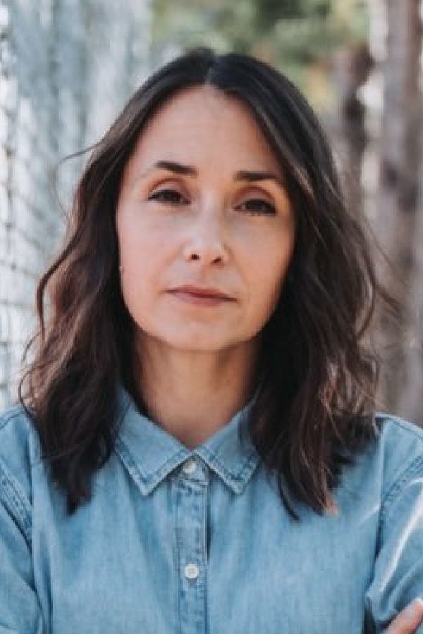 Person in light blue denim shirt standing against a brick wall, looking thoughtfully at camera.