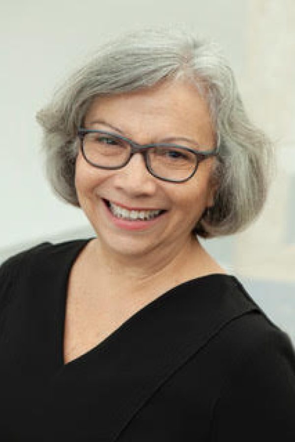 A person with short gray hair and glasses wearing a black top, smiling at the camera against a light background.