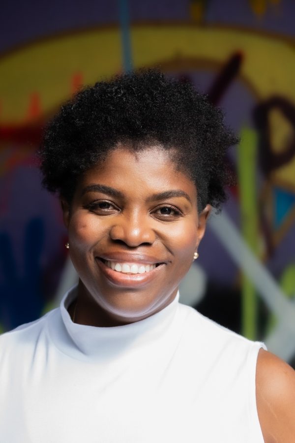 A smiling person wearing a white sleeveless shirt stands in front of a colorful, blurred graffiti wall background.