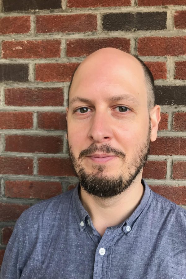 Person with beard wearing blue button-up shirt, standing in front of red brick wall.