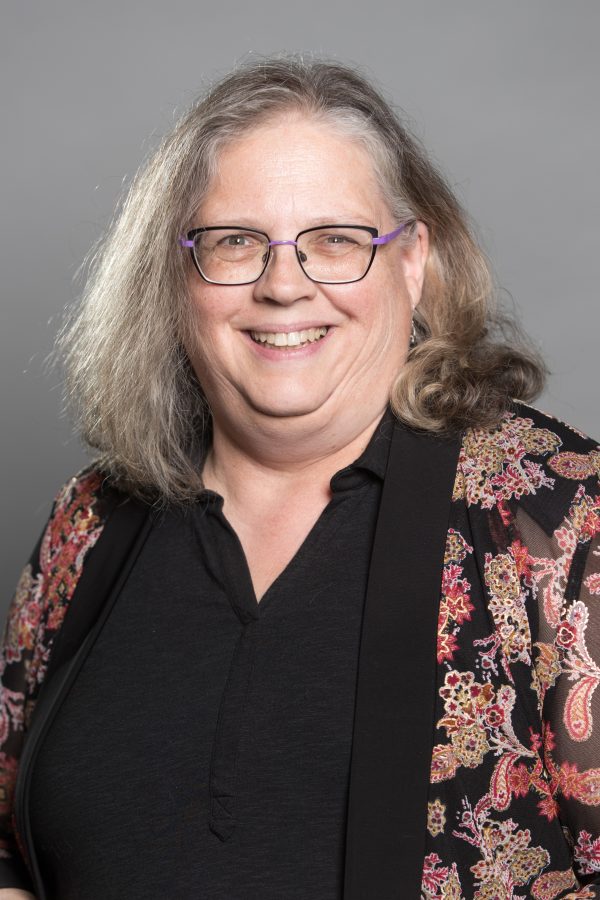 Person with long gray hair wearing glasses and a black shirt with a floral patterned cardigan, smiling at camera.