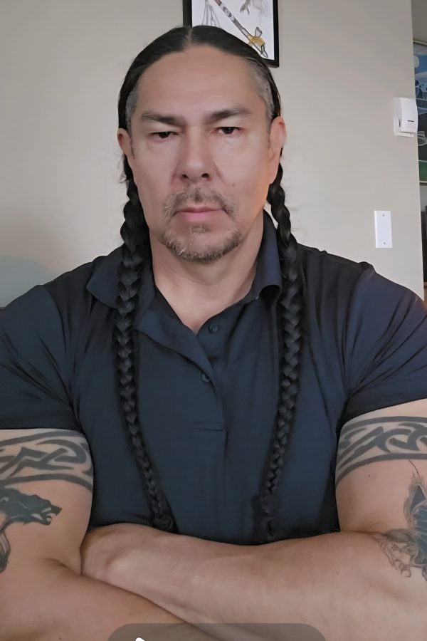 Person with long braided black hair and tribal arm tattoos wearing a dark blue shirt, arms crossed, indoors.