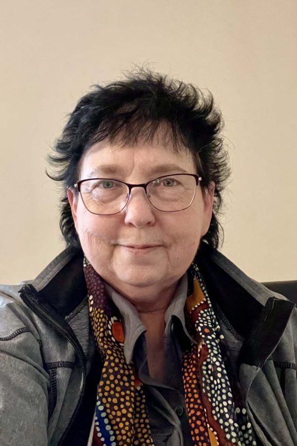 Person wearing glasses and a dark jacket over a colorful patterned shirt, smiling at the camera against a neutral background.