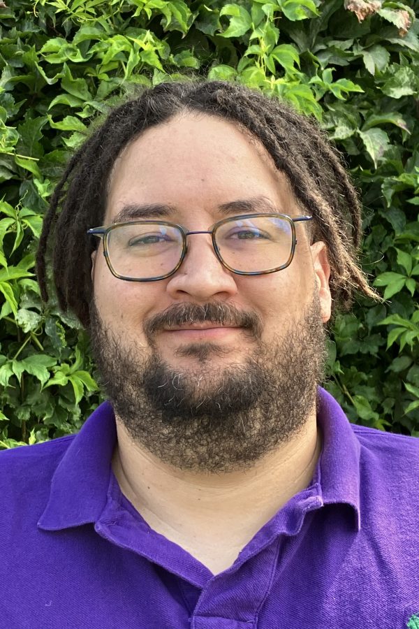 Person with dreadlocks and glasses wearing a purple polo shirt, photographed against a green leafy background.