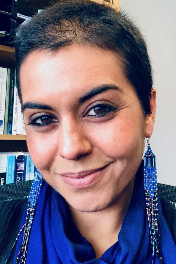 Person with short dark hair wearing a blue scarf and beaded earrings, smiling at the camera with bookshelves in the background.