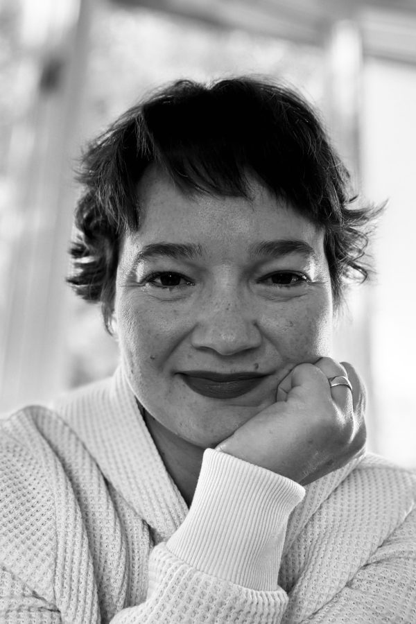 Black and white portrait of a person with short dark hair, wearing a light-colored ribbed sweater, resting their chin on their hand and smiling at the camera.