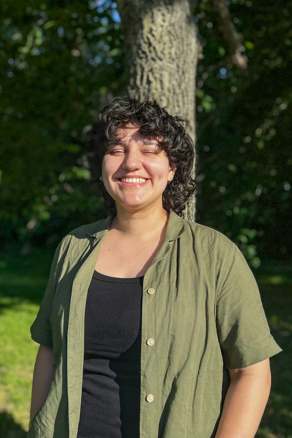 A smiling person with curly dark hair wearing a sage green button-up shirt over a black top, standing outdoors with trees in the background.