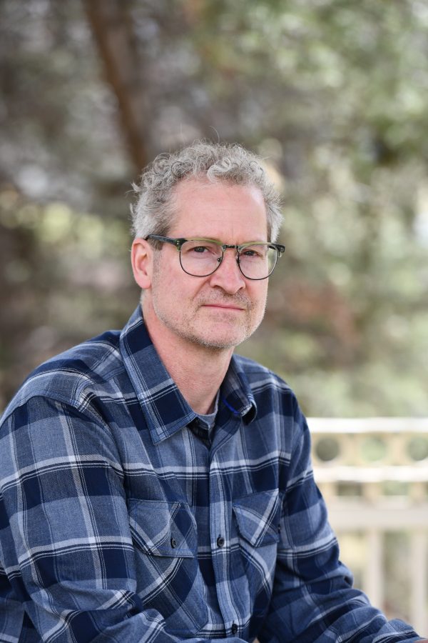 Person with gray hair and glasses wearing a blue plaid shirt, smiling outdoors with blurred foliage background.