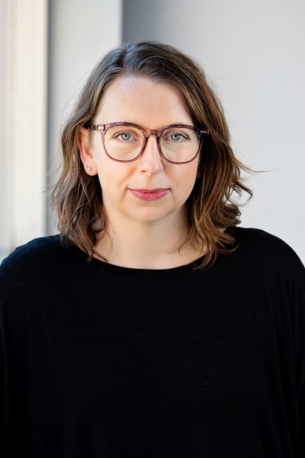 Person with shoulder-length brown hair and glasses wearing a black shirt, smiling at the camera against a blurred white background.