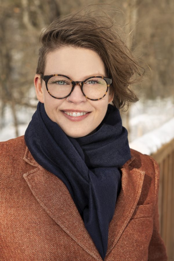 Person wearing glasses and a rust-colored blazer with a dark scarf, smiling at the camera outdoors in winter.