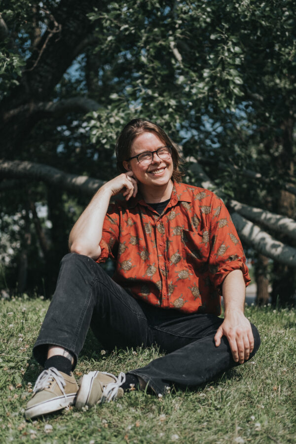 Person wearing glasses and an orange patterned shirt sitting on grass beneath leafy trees, smiling at the camera.