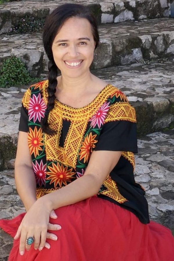 Person smiling at camera wearing colorful patterned top and red skirt, seated against stone steps.