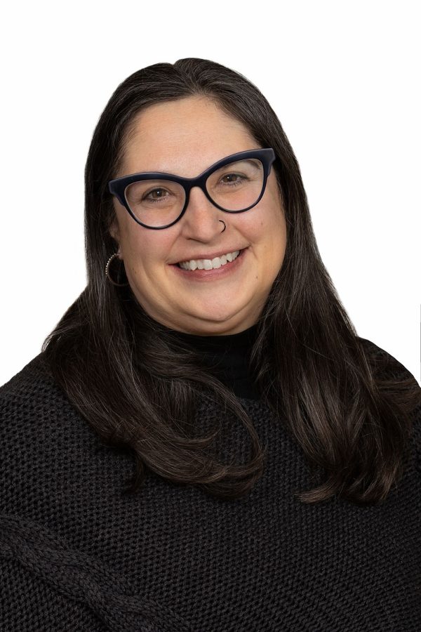 Person with long dark hair and glasses wearing a black textured sweater, smiling at the camera against a white background.