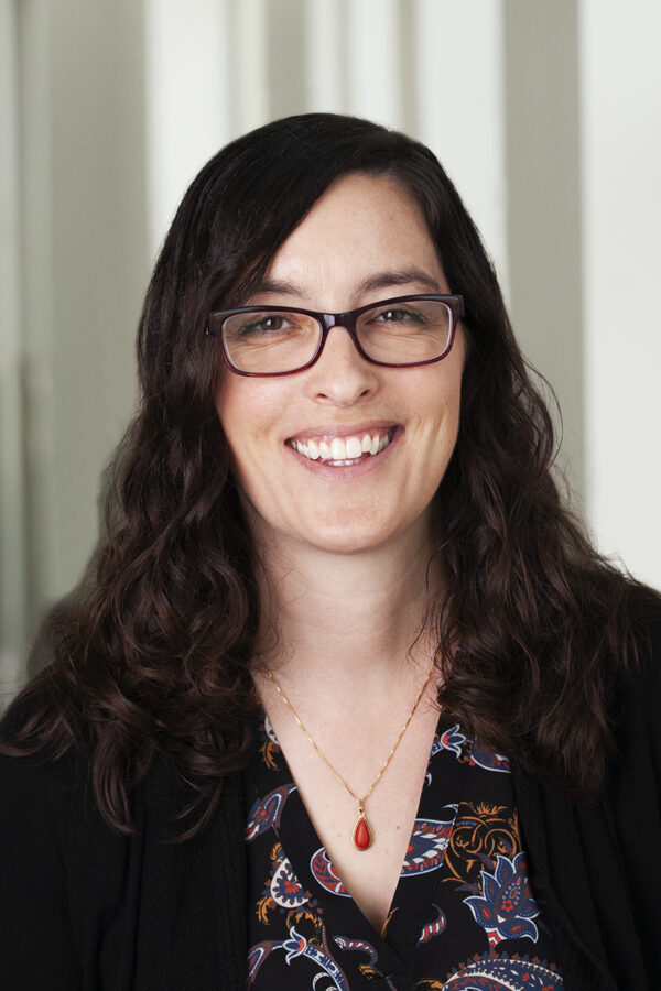 Person with long dark hair and glasses smiling at camera, wearing black cardigan and patterned top with red pendant necklace.