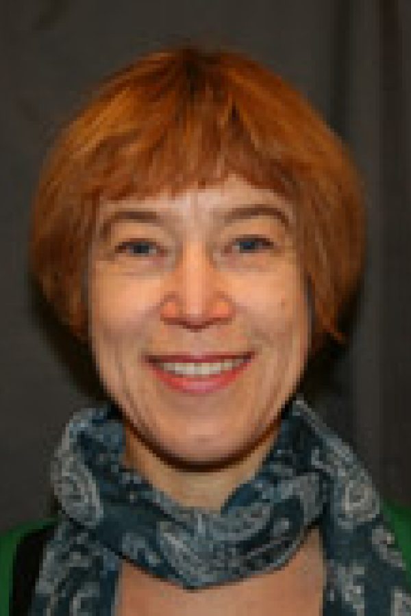 Person with short reddish-orange hair wearing a green top and patterned scarf, smiling at the camera against a dark background.