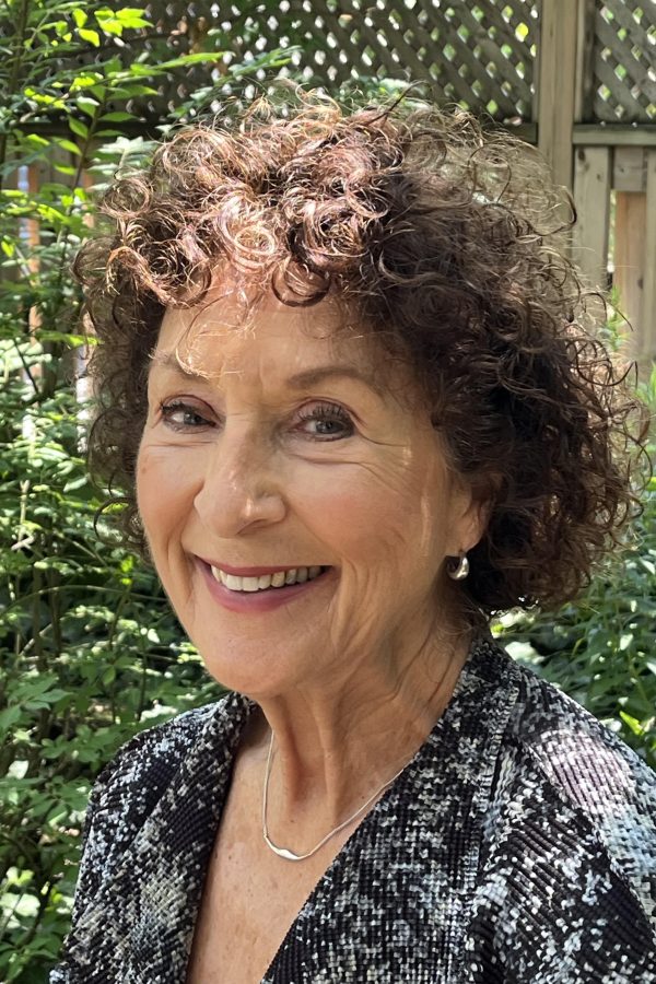Smiling person with curly gray hair wearing a dark sequined top and silver necklace, outdoors with greenery and lattice fence in background.