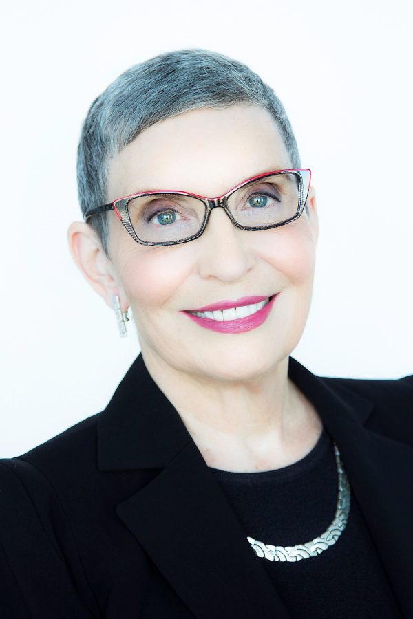 Person wearing glasses and pink lipstick, black blazer and necklace, smiling at camera against white background.