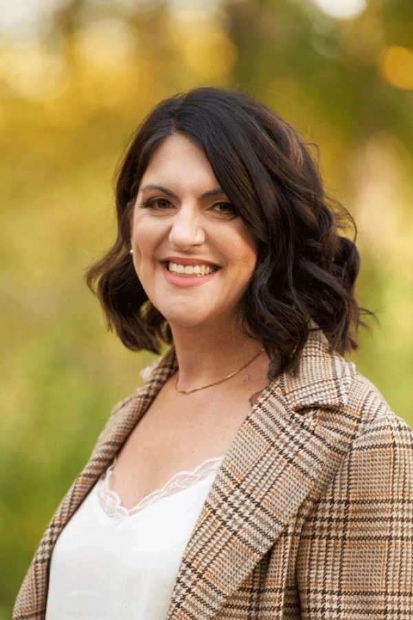 Person with dark wavy hair smiling at camera, wearing a tan plaid blazer and white top, with blurred green foliage background.