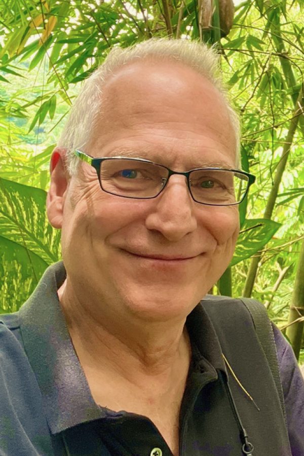 Smiling person with white hair and glasses wearing a dark jacket against a background of green bamboo foliage.