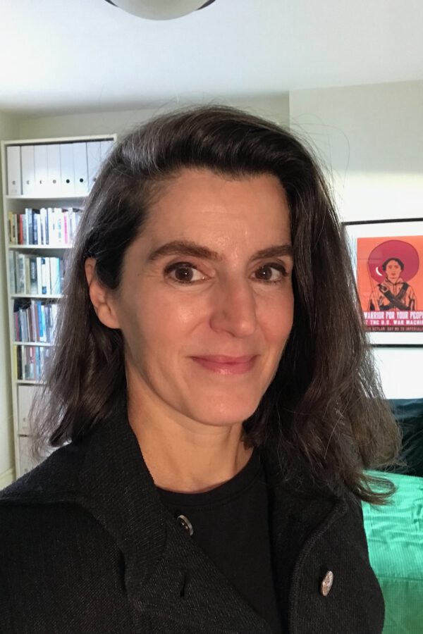 Person with long dark hair wearing a black blazer, smiling at the camera in a home office with bookshelves and artwork visible behind them.