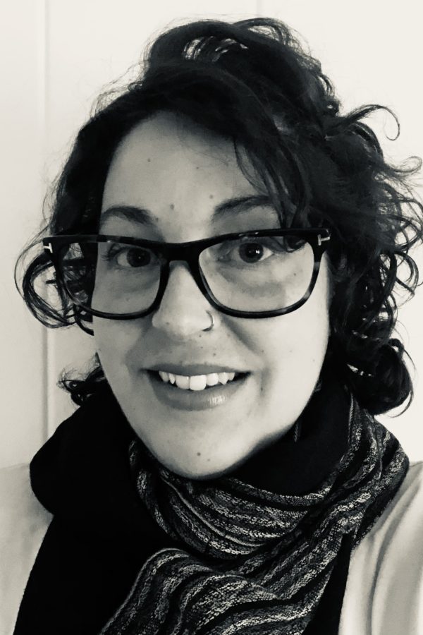 Person with black curly hair and glasses wearing a black scarf, smiling at the camera in a black and white portrait.