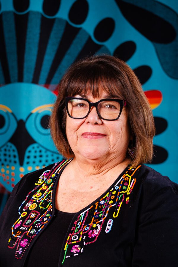 Person wearing glasses and a colorful beaded vest against a vibrant blue and black geometric patterned background.