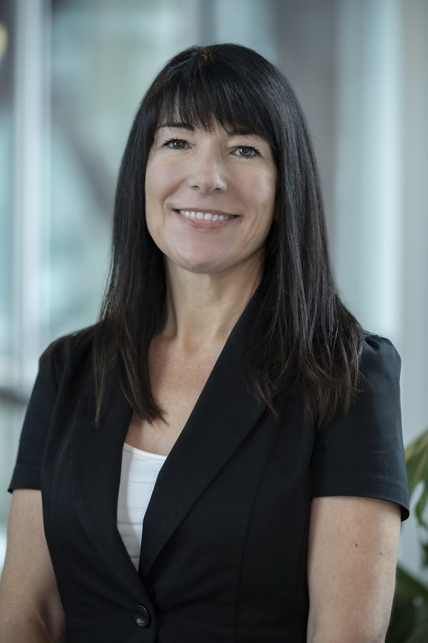Professional headshot of a person with long dark hair and bangs, wearing a black blazer over a white top, smiling at the camera in an office setting.