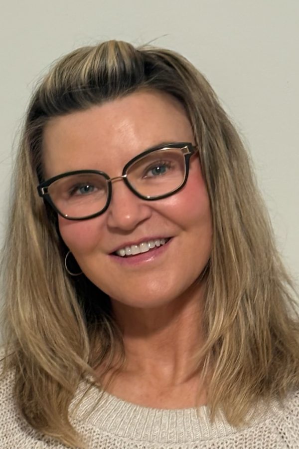 Person wearing black-framed glasses and a light sweater, smiling at the camera against a neutral background.