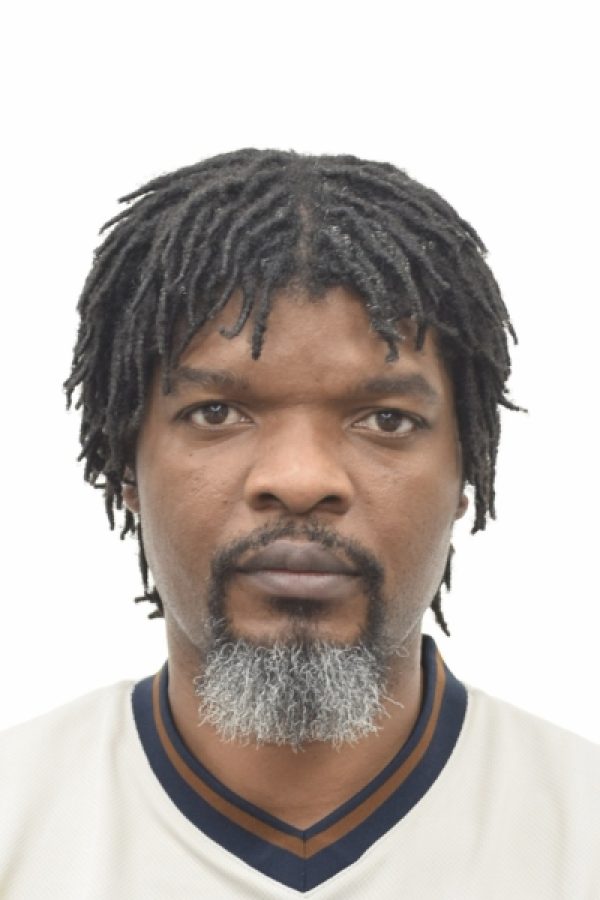 Portrait of a person with dreadlocks and gray beard wearing a white v-neck shirt with dark trim against a white background.