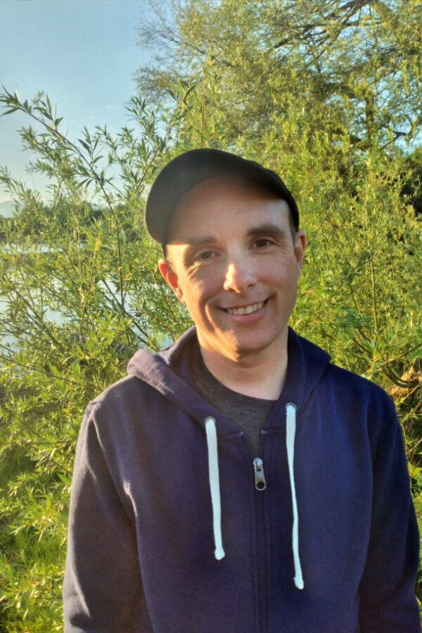 Person wearing dark hoodie and cap smiling outdoors surrounded by green foliage and trees on a sunny day.