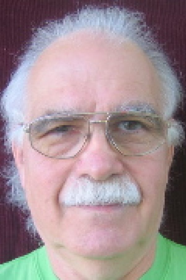 Older person with white hair and glasses wearing a green shirt, photographed against a burgundy background.