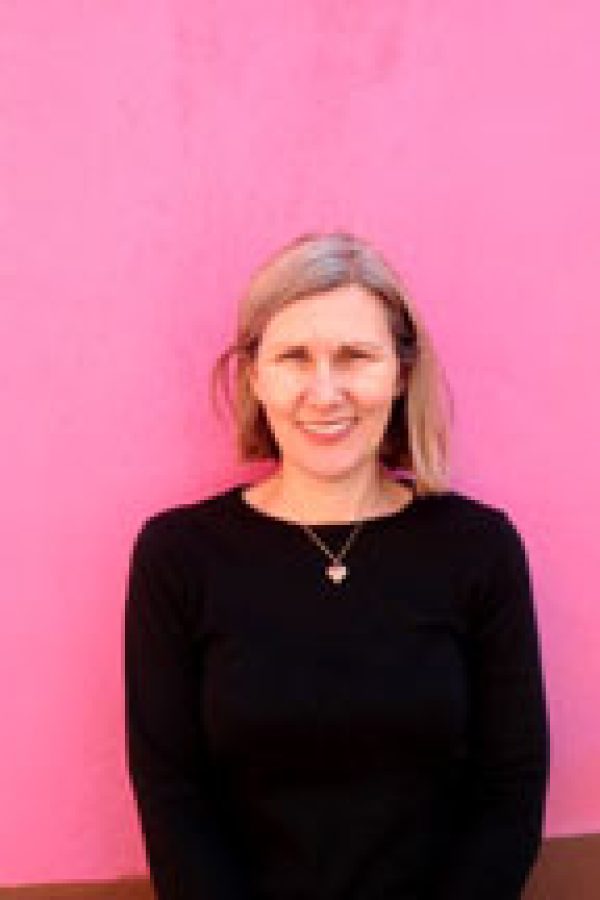 Person with blonde hair wearing a black sweater and cross necklace against a bright pink background.