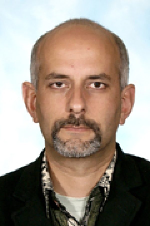 Portrait of an adult person with short hair and facial hair, wearing a dark jacket over a collared shirt, against a light background.