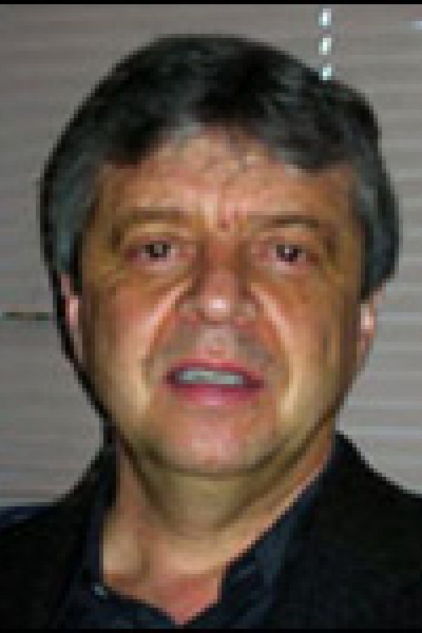 Headshot of a person with dark hair and facial hair wearing a dark colored shirt against a gray background.