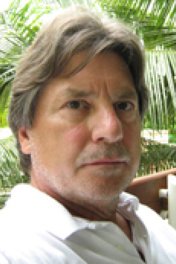 Person with shoulder-length hair smoking a cigar, wearing a white shirt, with palm trees in the background.