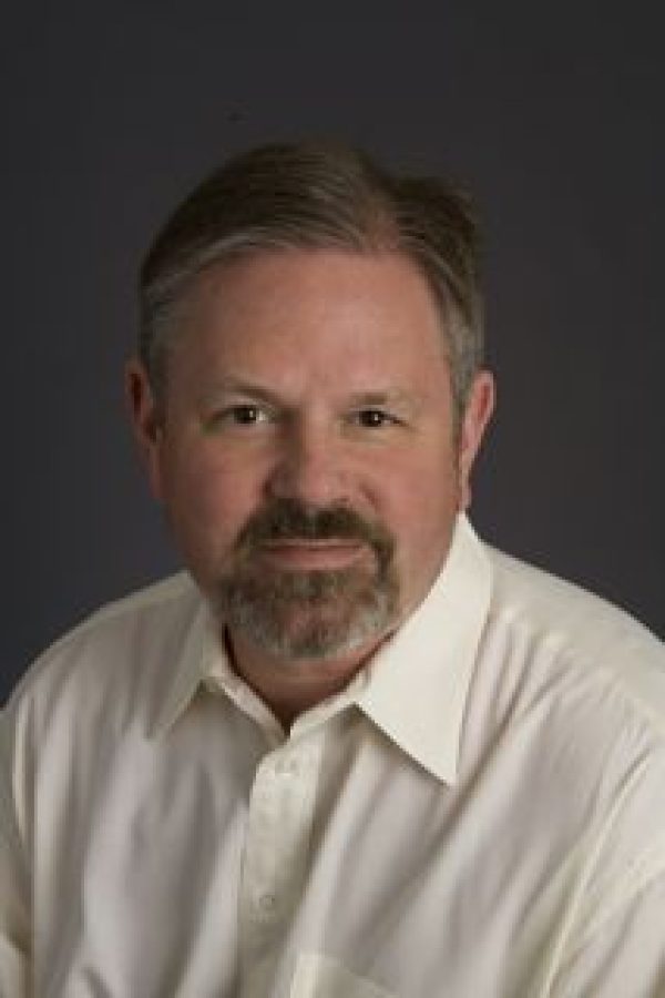 Professional headshot of a person with short brown hair and facial hair, wearing a light-colored collared shirt against a gray background.
