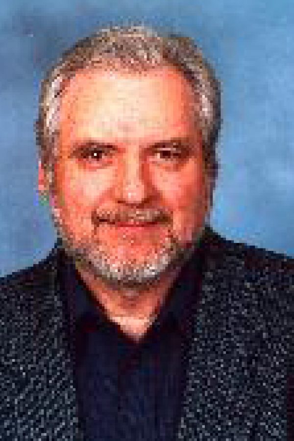 Portrait of an older person with gray hair and mustache wearing a dark sweater against a blue background.